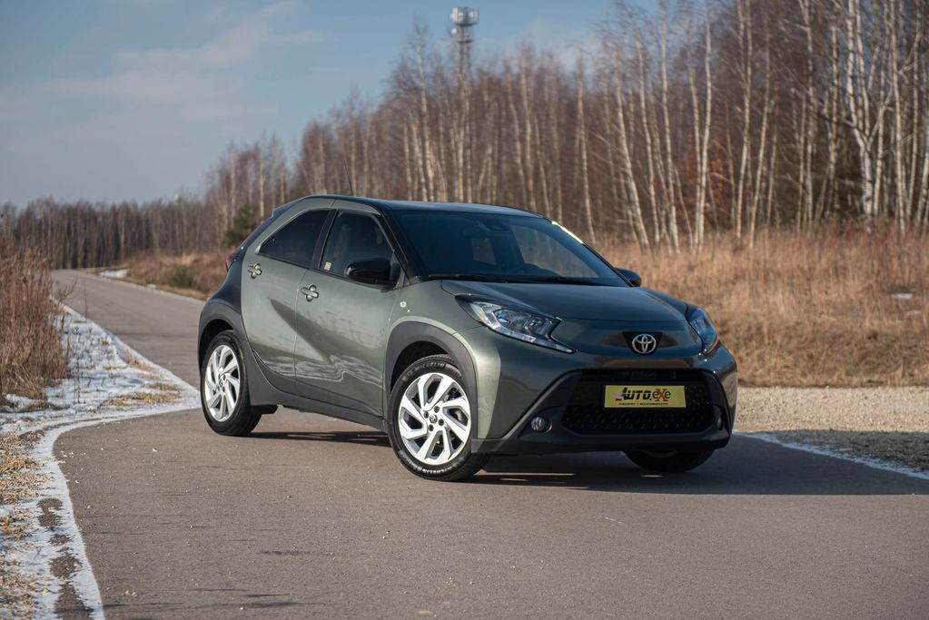 Toyota Aygo X STYLE grafit/czarny stoi prawym przednim bokiem na drodze asfaltowej obok traw