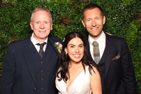 Bride and two men in suits smiling in front of botanical backdrop