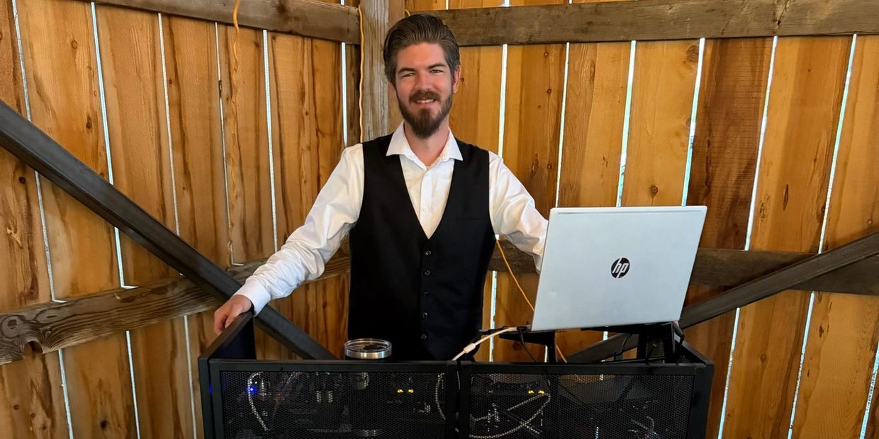 Man in vest standing behind DJ booth with laptop in wooden room.