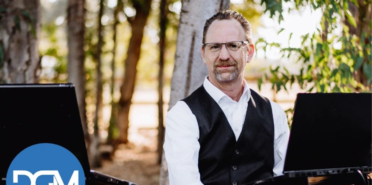 Man in glasses and vest sitting outdoors between two laptops.