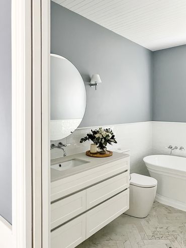 Light-filled Hamptons bathroom with herringbone marble floors, shaker cabinetry & round mirror