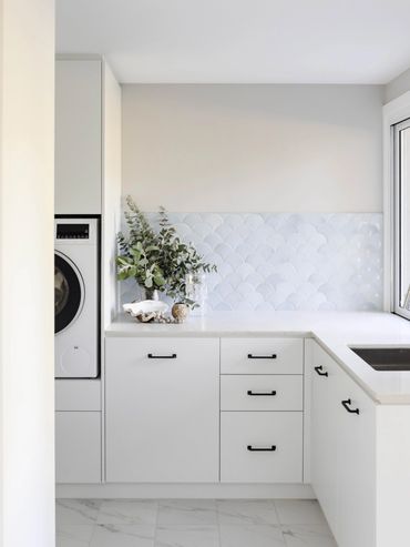Light-filled coastal laundry with marble floor, white cabinetry, black handles and blue fish-scale t