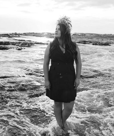 Woman in black dress with feather headpiece standing in the ocean surf.