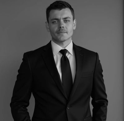 Confident man in a black suit and tie against a plain background.