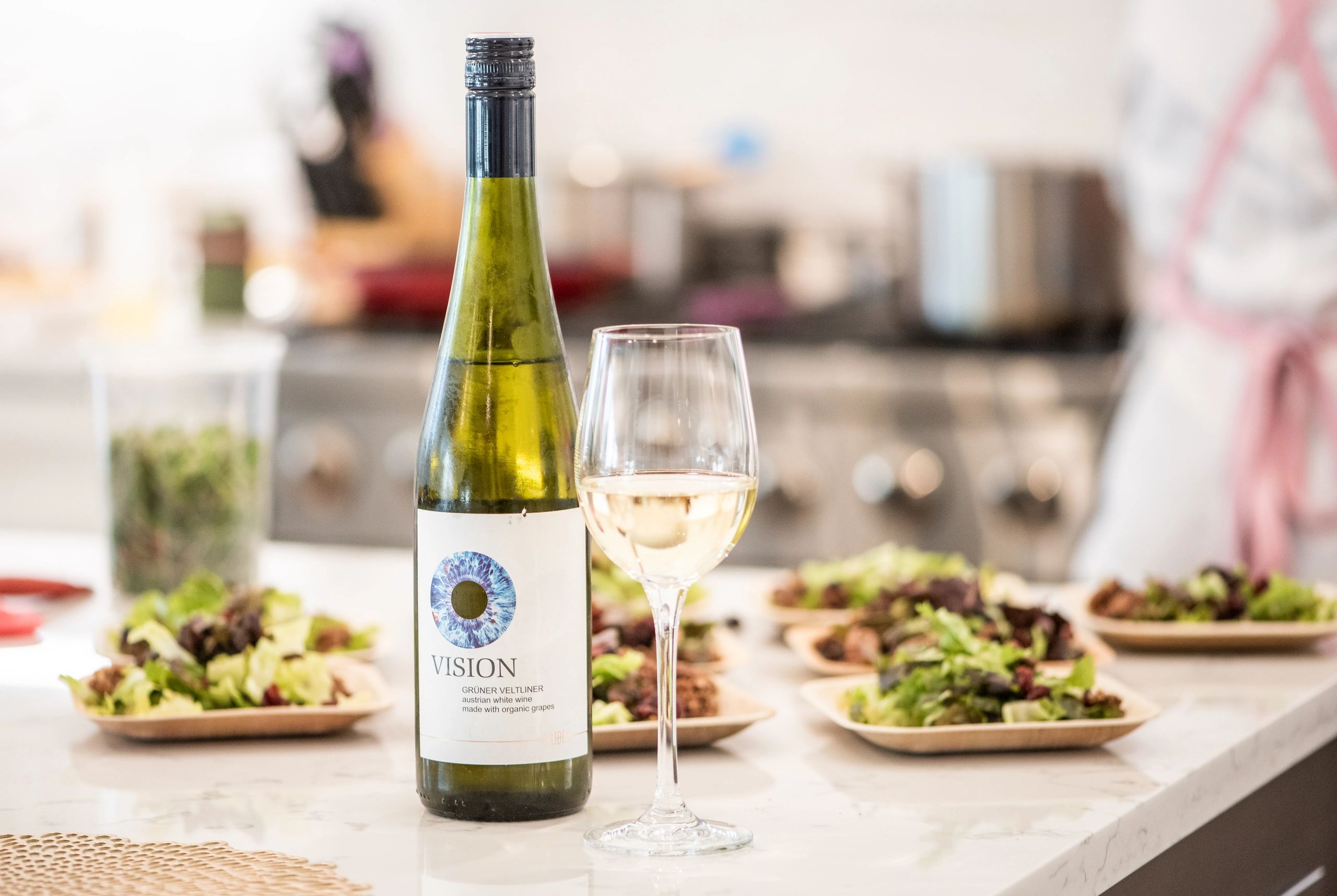 A bottle of wine and a glass of white wine sitting in a kitchen in front of a row of salad plates.