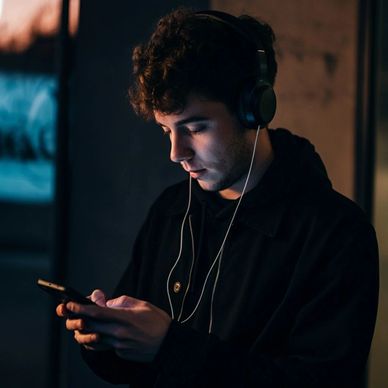 Young man wearing headphones, focused on his phone in dim light.