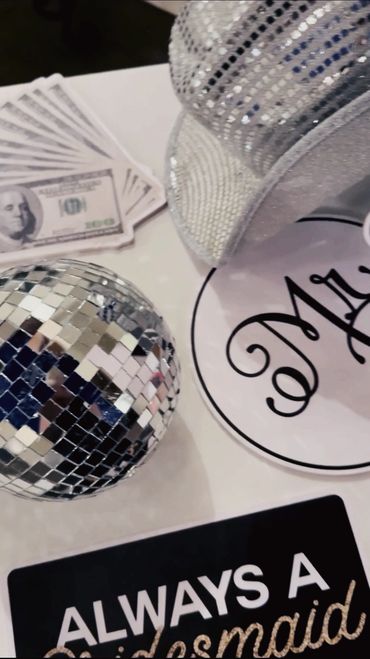 Disco ball, money, glittery hat, and a bridesmaid sign on a table.