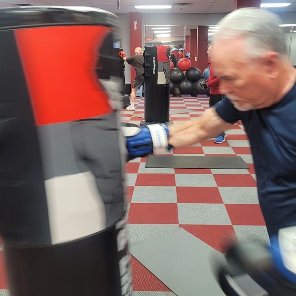 Older man boxing a punching bag in a gym with others training in the background.