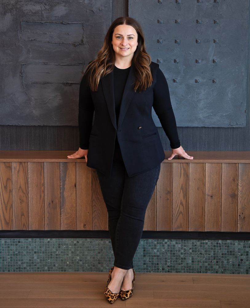 A woman in a black outfit and leopard print heels stands confidently indoors.