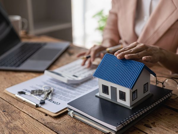 A person's hands on a small model of a house, with a calculator and papers on a desk.