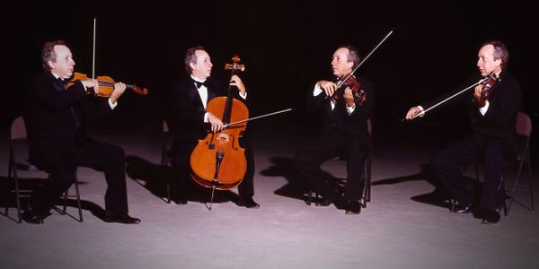 Four musicians in tuxedos play string instruments in a dark setting.