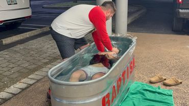 A man baptizes another person in a metal tub filled with water outdoors at night.