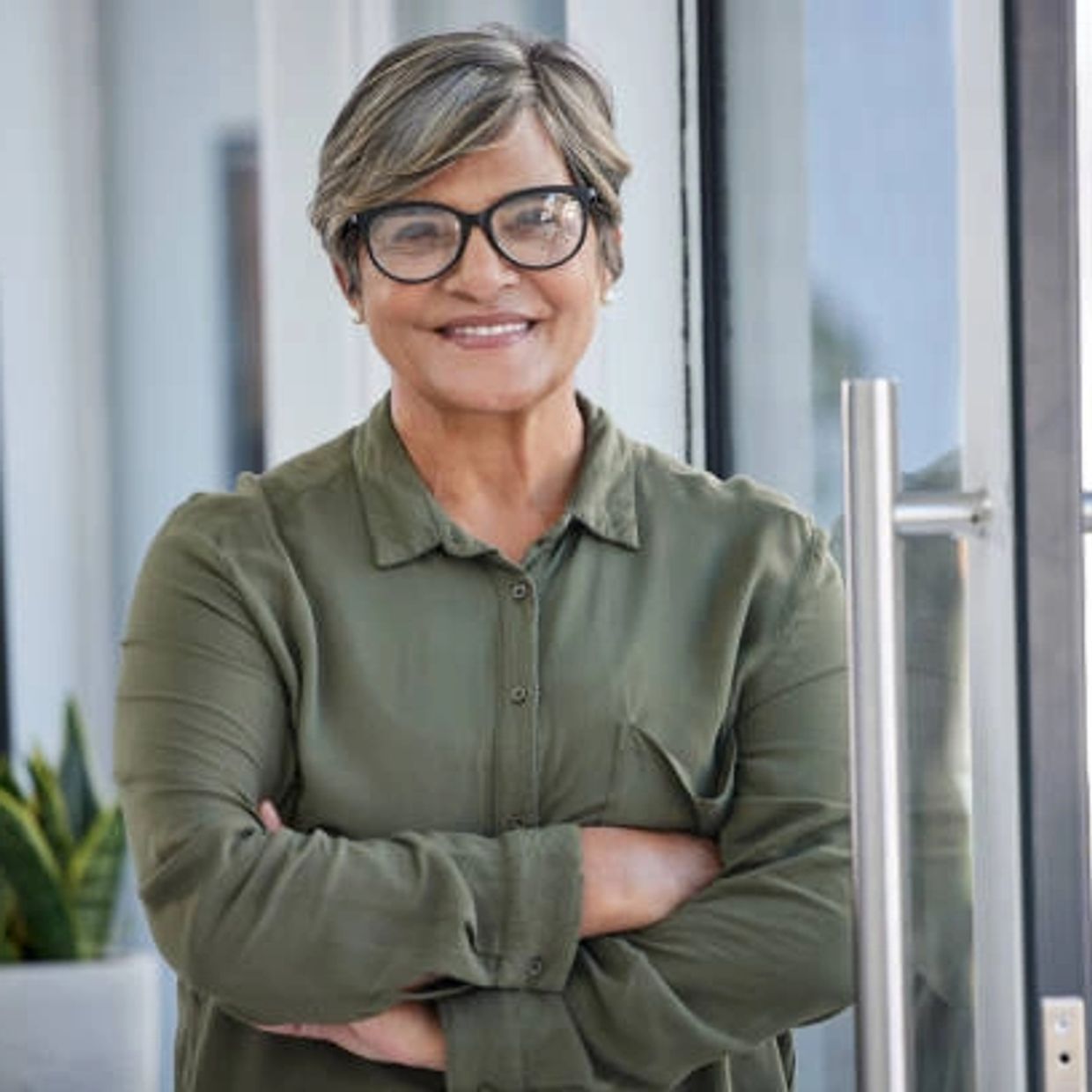 Confident woman with glasses smiles, standing with arms crossed indoors.