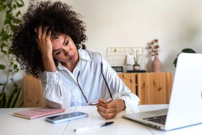 Tired woman working at a desk with laptop and phone.