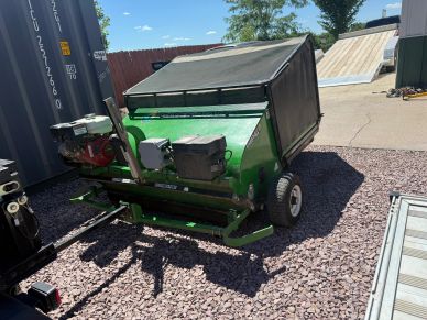 Green turf maintenance machine parked on gravel under sunlight.