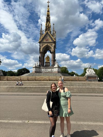 Two young women posing in front of a tall, ornate monument under a partly cloudy sky.