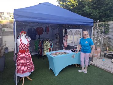 Woman stands beside a market stall with vintage clothing and accessories.