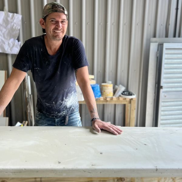 man in his workshop with t-shirt and cap standing behind a concrete work surface, smiling at the cam