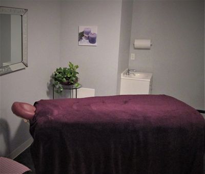 Body treatment room has a massage table with purple lines. Glass table, plant, sink, & spa picture