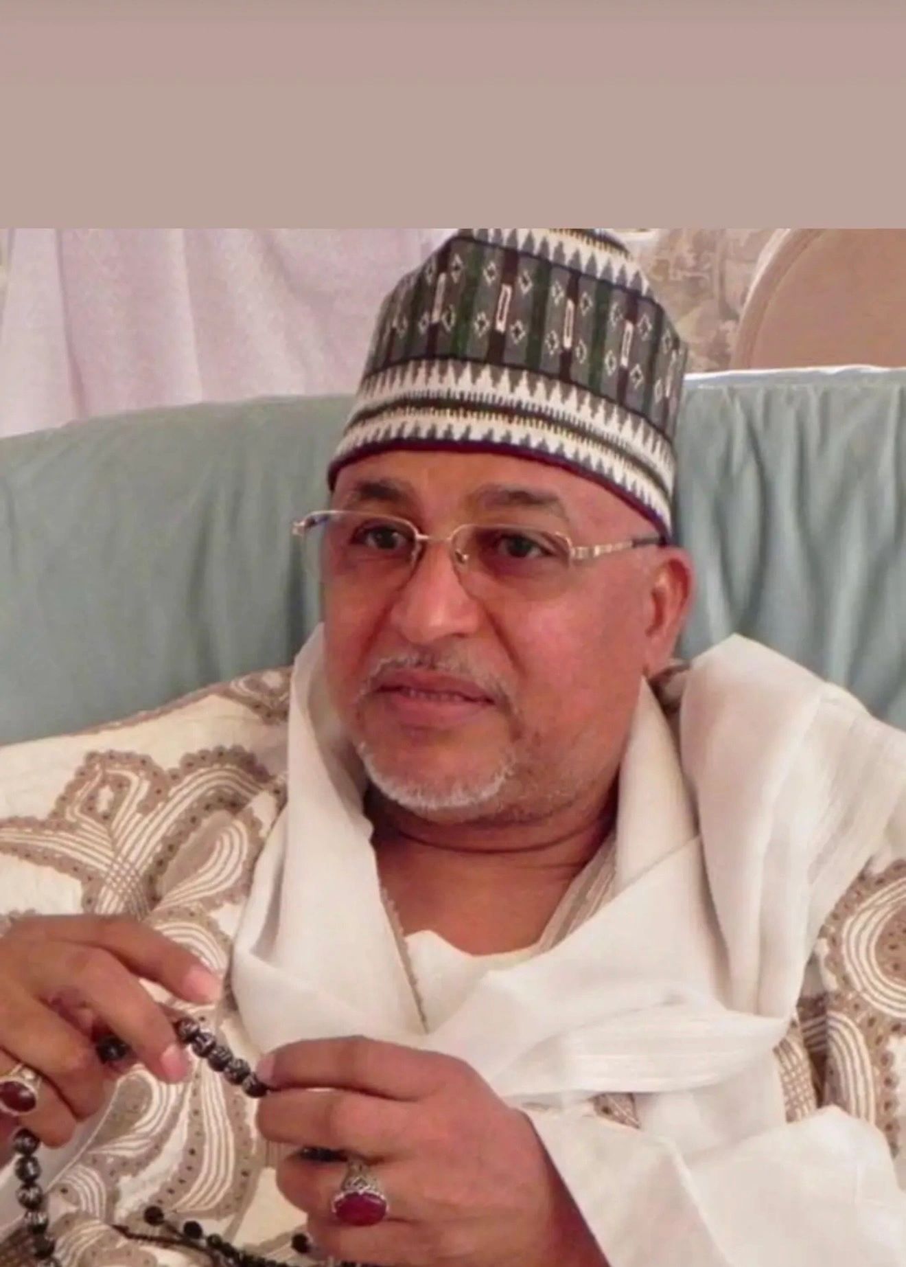 Elderly man wearing traditional attire and a patterned cap holding prayer beads.