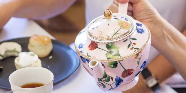 Hand holding a floral teapot over a tea cup and plate.