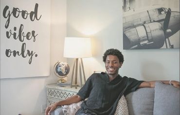 A man smiles sitting on a couch with a 'good vibes only' sign behind him.