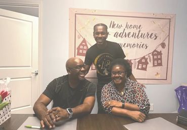 Three people smiling in a room with a 'New home adventures memories' banner.