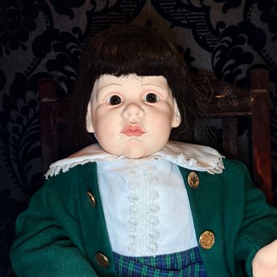 A vintage doll with a porcelain face and dark hair wearing a green jacket and white lace collar.