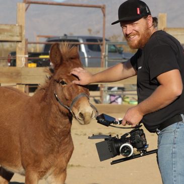 Man smiling while petting a small horse holding a camera.