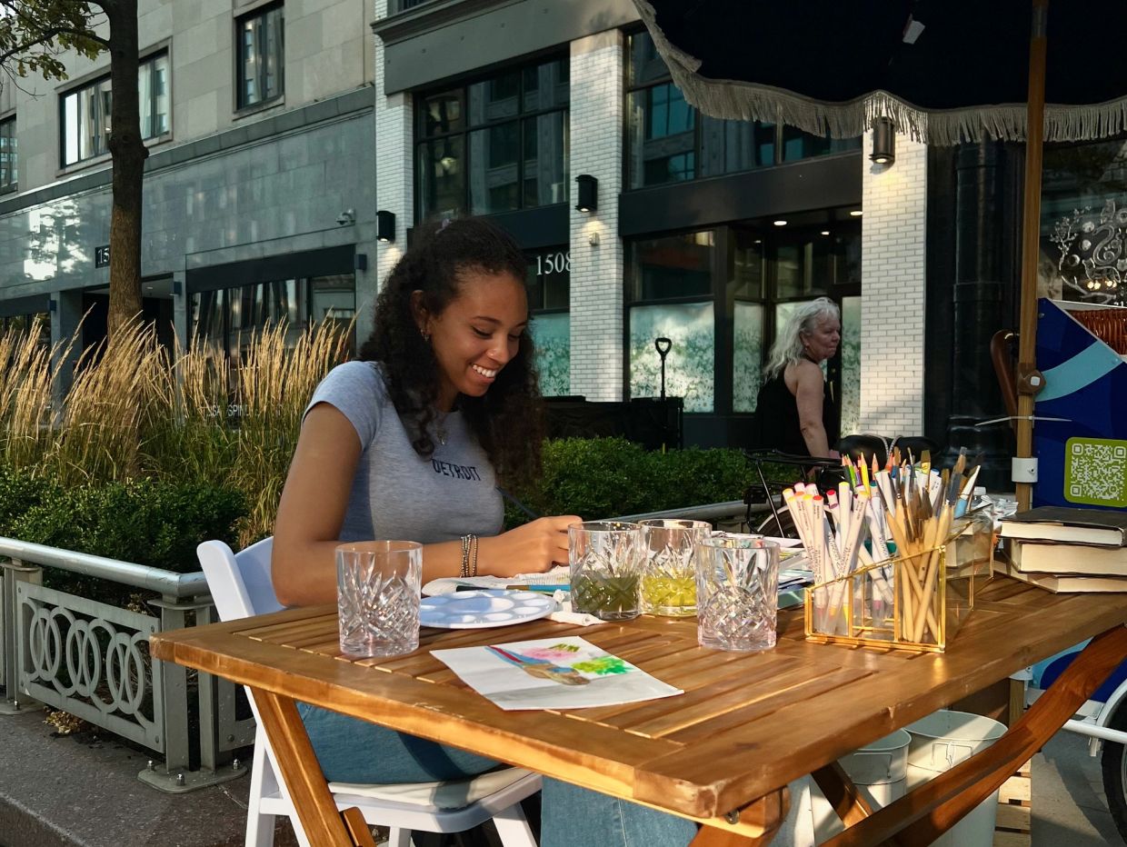 A woman happily painting at an outdoor wooden table with art supplies.