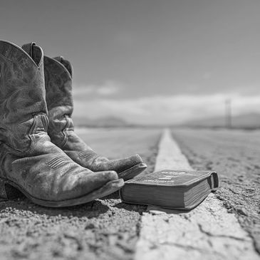 Worn cowboy boots and a book on a deserted road under a clear sky.