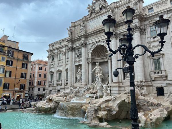 View of the Trevi Fountain with buildings and a street lamp under a cloudy sky.