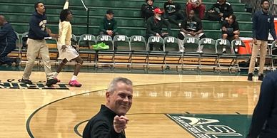 Man pointing and smiling on a basketball court with players and spectators in the background.