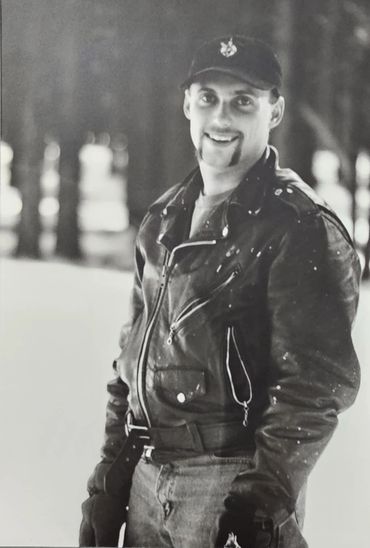 Smiling man in leather jacket and cap with paint splatters.