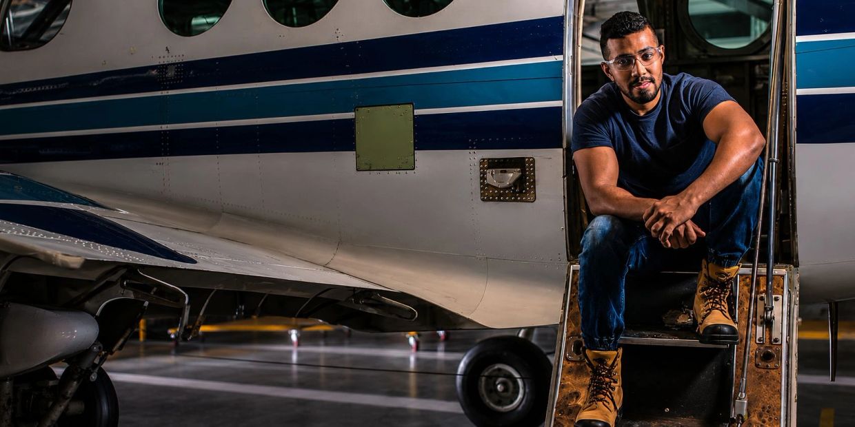 Man sitting on airplane stairs in a hangar, wearing boots and safety glasses.