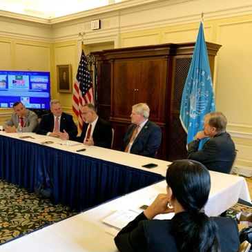 Officials seated at a conference table during a formal meeting with flags in the background.