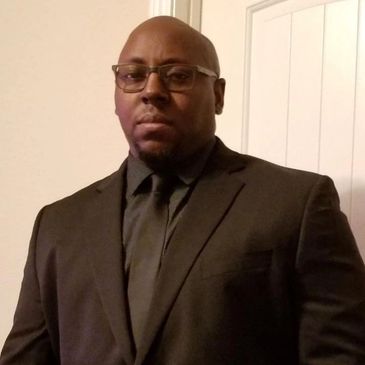 Man wearing a black suit and glasses posing indoors.
