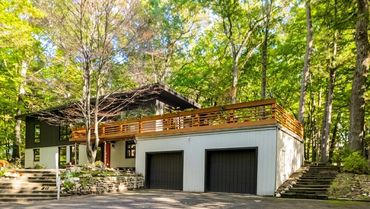 Modern house nestled among lush green trees with a spacious garage and wooden balcony.