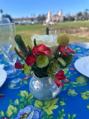 A small flower arrangement in a white vase on a blue floral tablecloth outdoors.