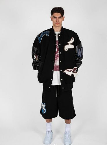 Young man in oversized black varsity jacket and shorts with patches, white sneakers.