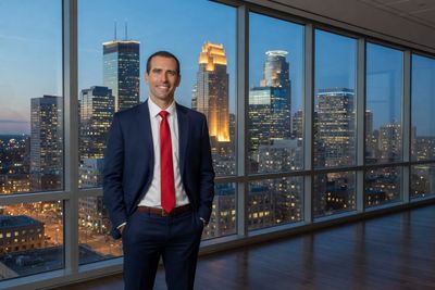 Man in a suit with a red tie stands in front of a city skyline at dusk.