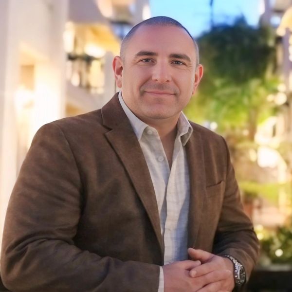 Man in brown blazer smiling outdoors in a relaxed setting.