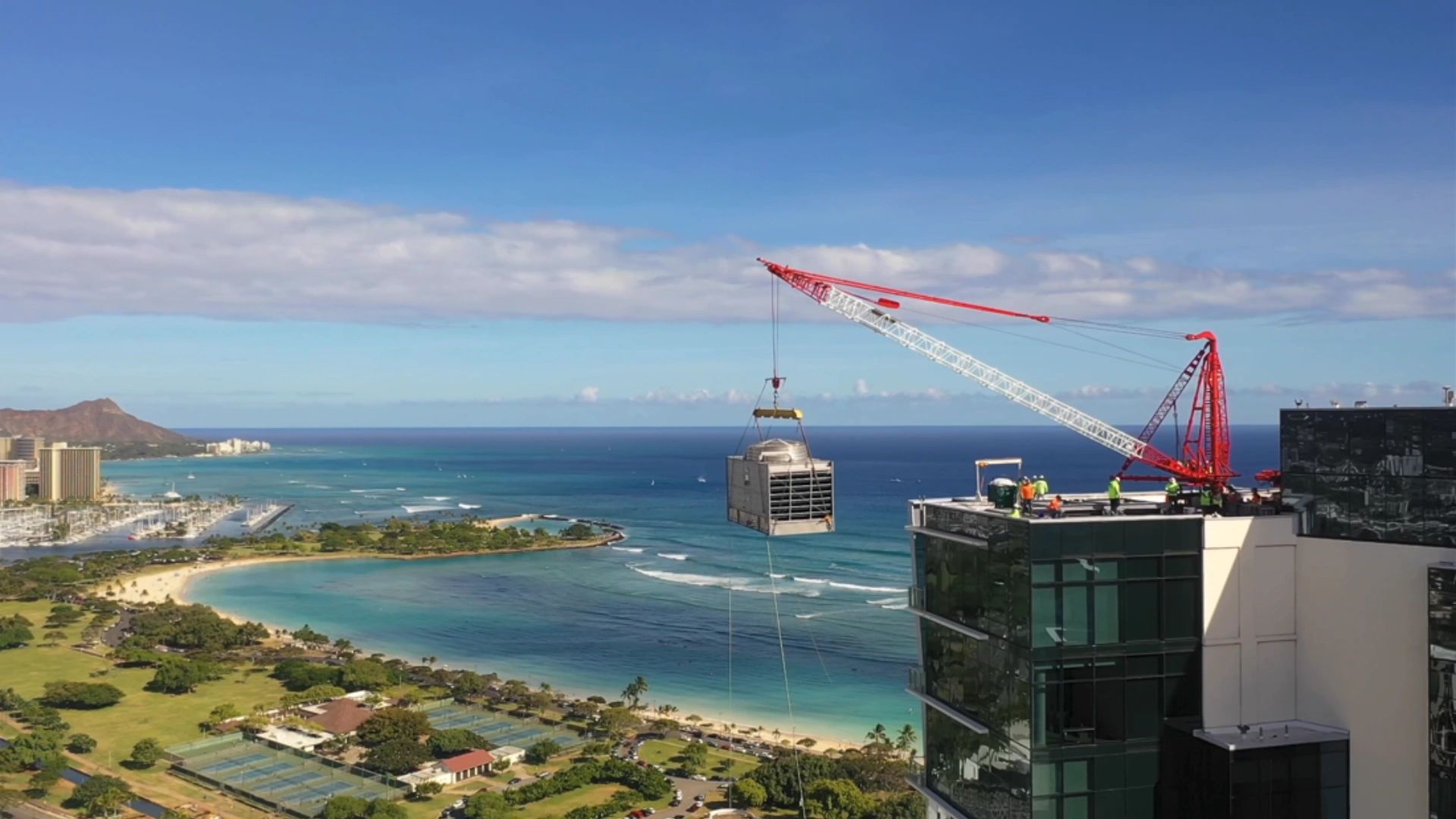 Crane lifting a large unit onto a building rooftop by the ocean.