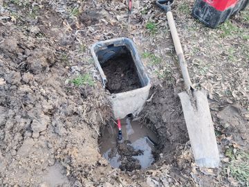 A shovel and a container in a muddy hole outdoors.
