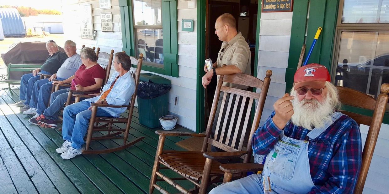 Photograph of 5 older people siting in rocking chairs on the porch of the airport terminal. 