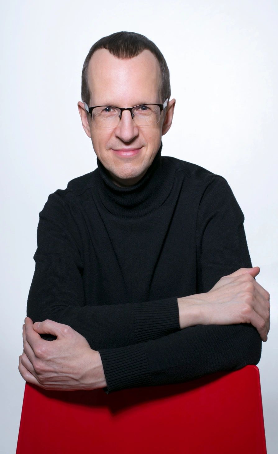Man in black turtleneck and glasses sitting with arms crossed on a red chair.