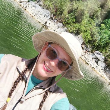 Mujer sonriente con sombrero y gafas de sol junto a un lago.