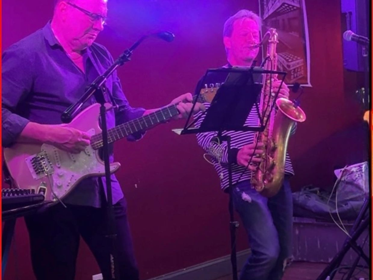 Two musicians playing guitar and saxophone on a purple-lit stage.