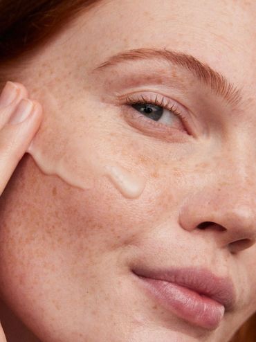 Close-up of a woman applying cream to her freckled cheek.