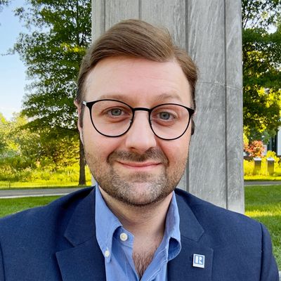 Man with glasses and beard smiling outdoors in business attire.